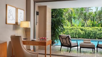 A hotel room with a desk, chair, and a view of a swimming pool surrounded by lush greenery.