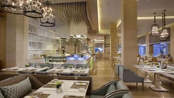 A spacious hotel dining area with multiple seating arrangements, including tables and booths, and a well-lit ambiance with hanging chandeliers.