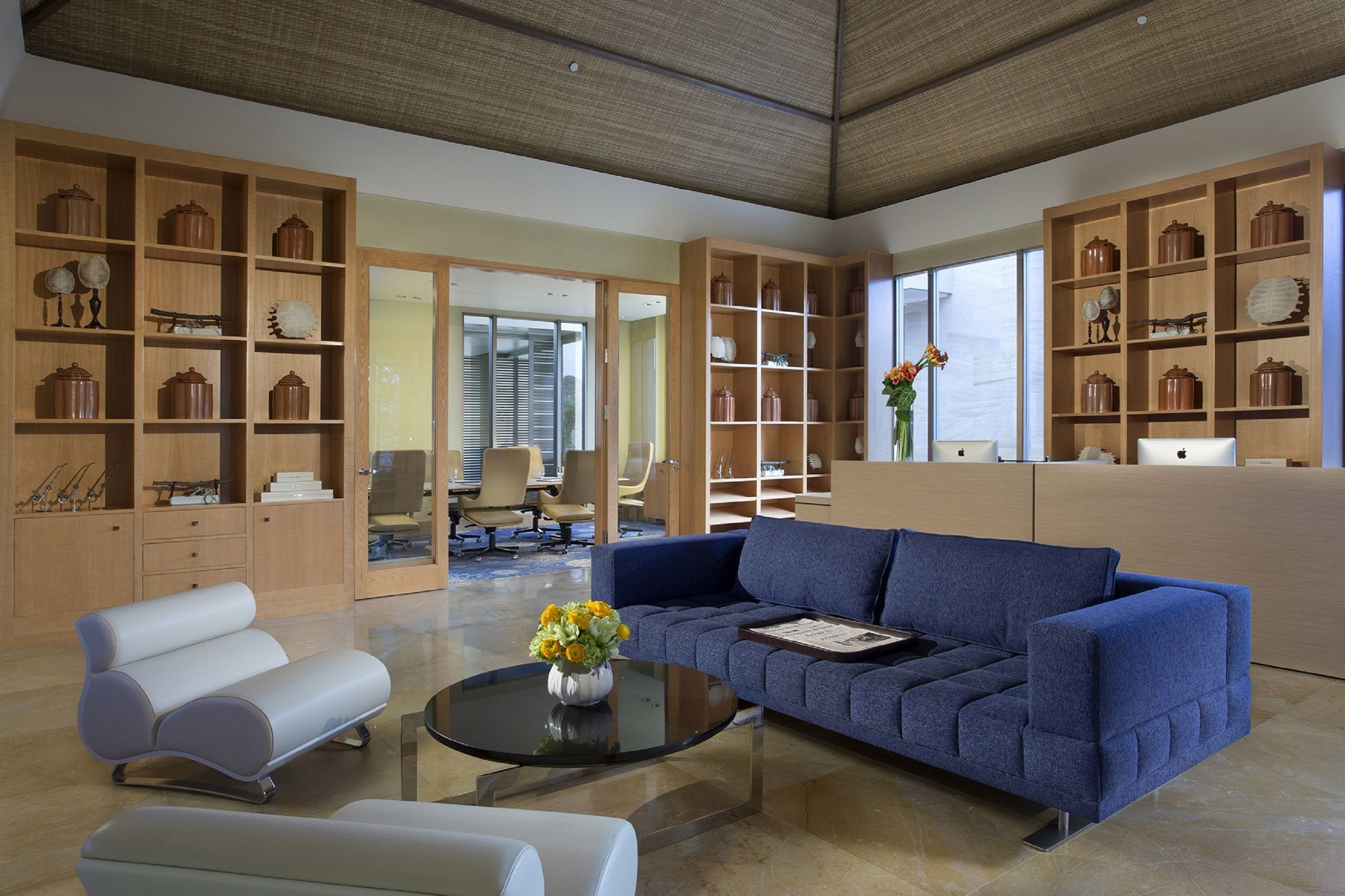 A modern living room with a blue sofa, white armchairs, and a glass coffee table. There are wooden bookshelves filled with decorative items and a large window providing natural light.