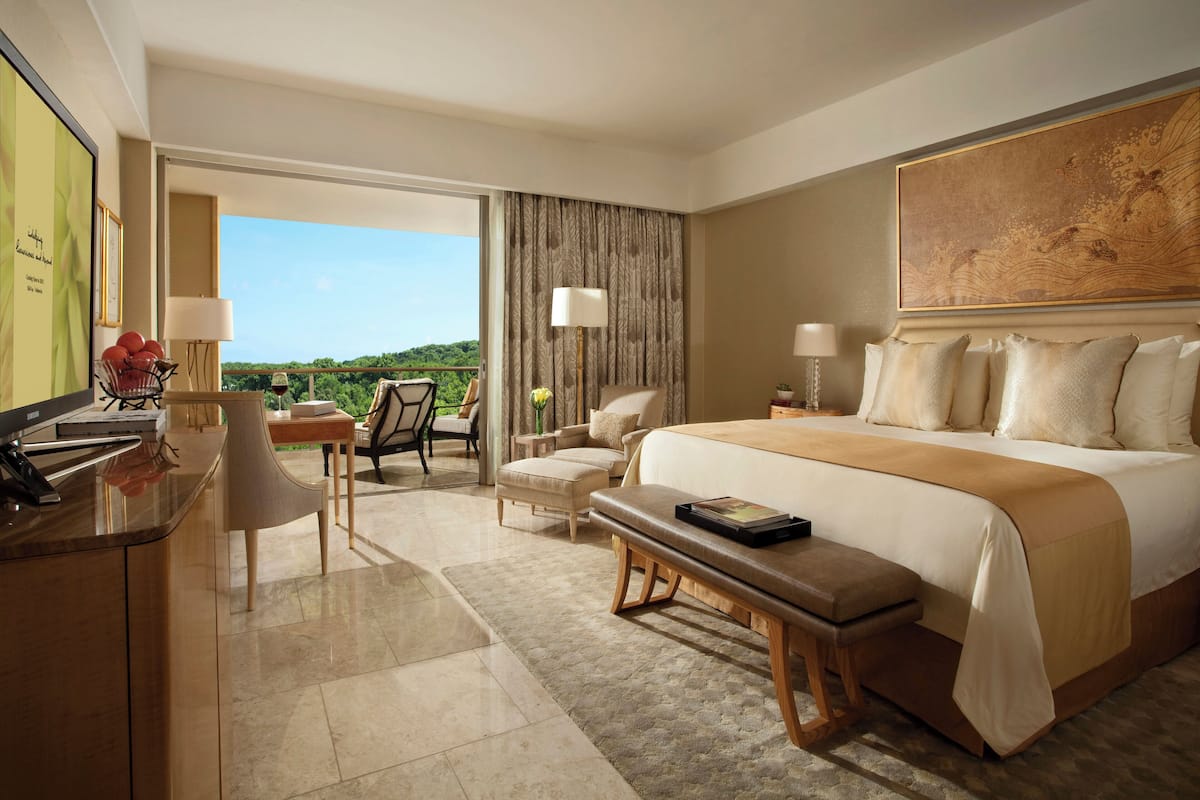 Habitación de hotel con una cama grande, un escritorio con computadora, una silla, una mesita con lámpara y vista al exterior.