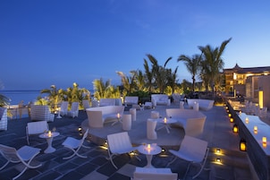 An outdoor seating area with tables and chairs, palm trees, and a view of the ocean.