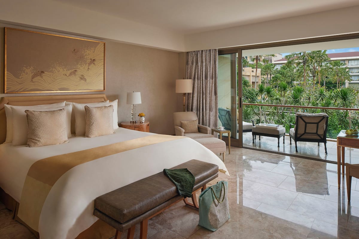 Habitación de hotel con una cama grande, un banco, una silla, una mesita y un balcón con vistas a palmeras y edificios.
