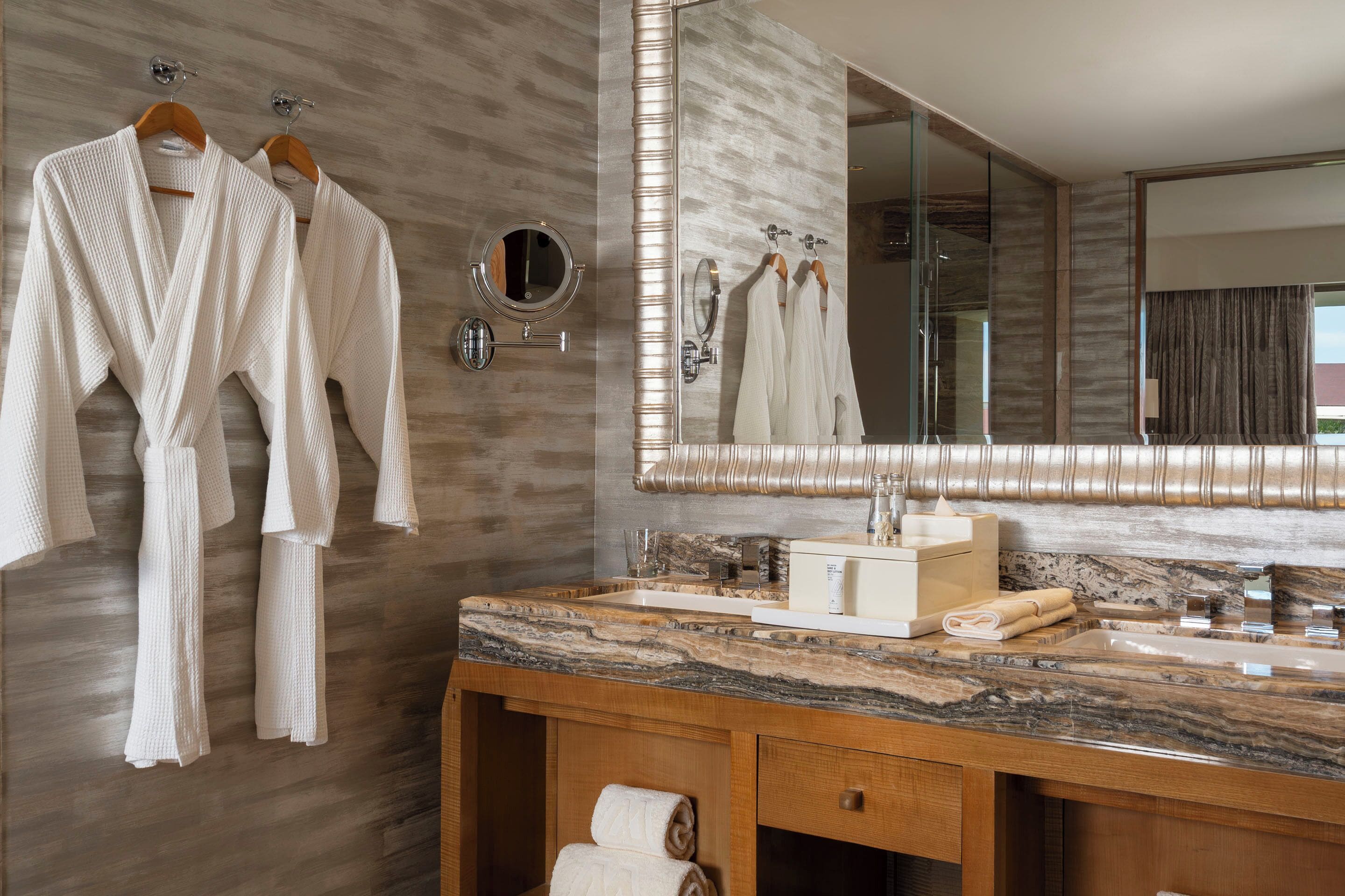 A modern bathroom with a marble sink, a large mirror, and two white robes hanging on wooden hangers.