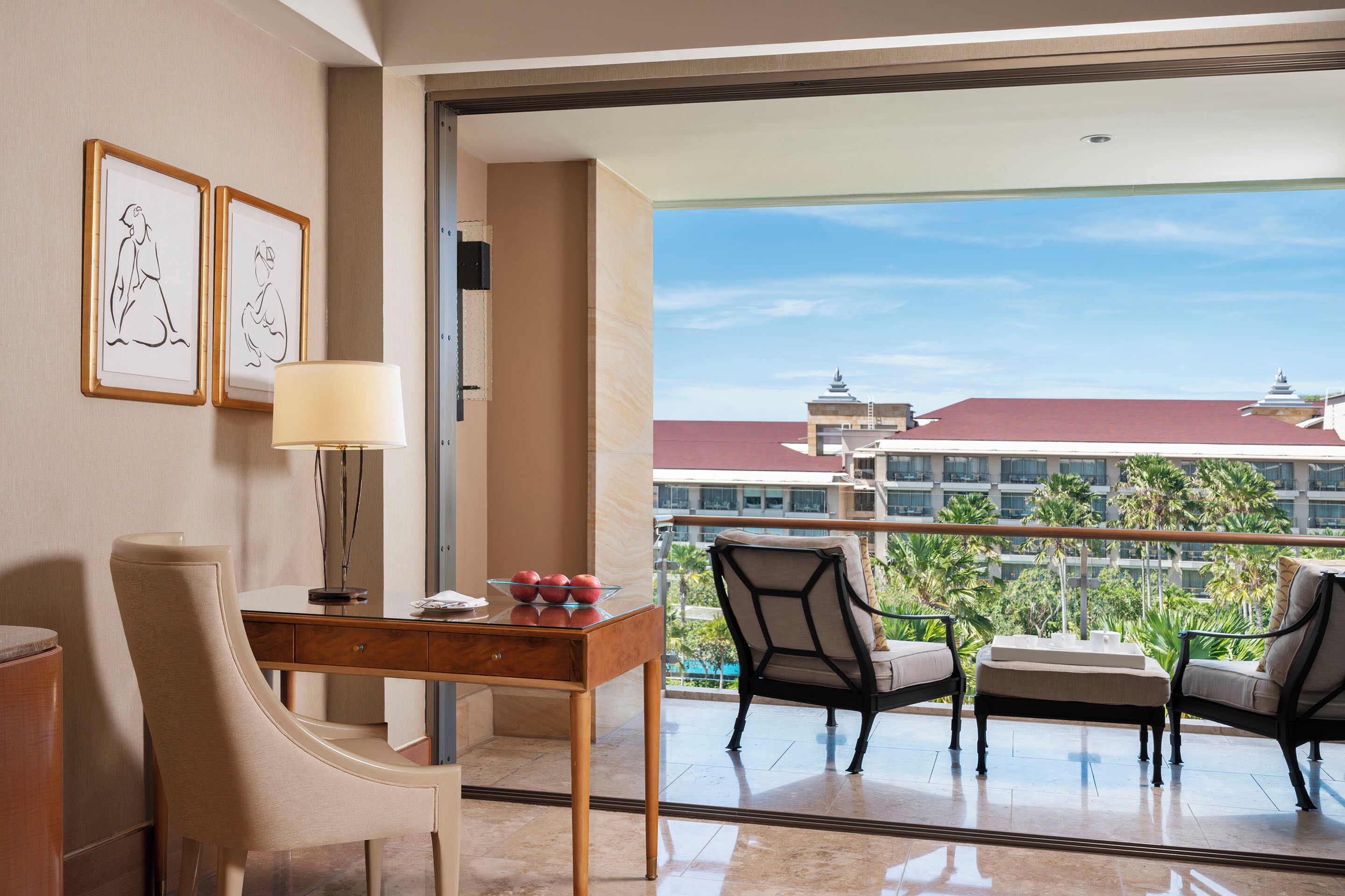 Habitación de hotel con escritorio, sillas, lámpara y vistas a un edificio y palmeras.