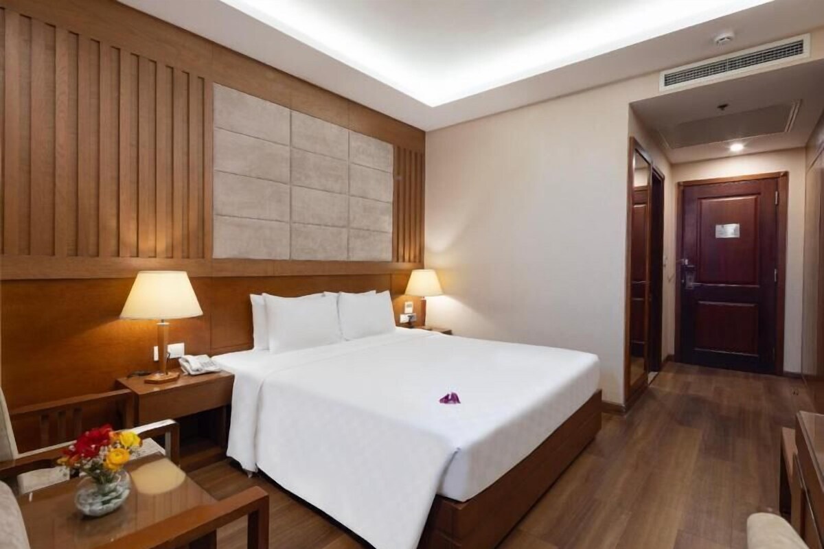 Une chambre d’hôtel avec un grand lit, une tête de lit en bois, deux tables de chevet avec des lampes, un petit bureau avec une chaise et un plancher en bois.