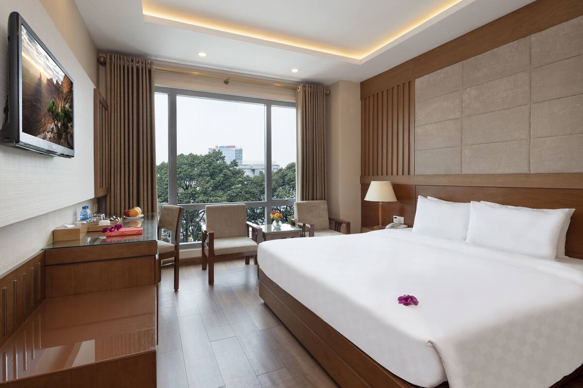 Une chambre d’hôtel avec un grand lit, un bureau, une chaise, une télévision et une vue sur les arbres et les bâtiments.