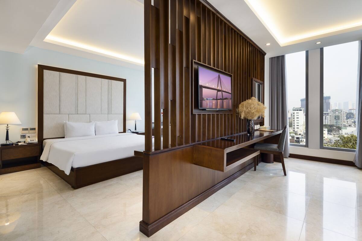 Une chambre d’hôtel moderne dotée d’un grand lit, d’un téléviseur fixé sur un panneau en bois, d’un bureau avec une chaise et offrant une vue sur la ville grâce à de grandes fenêtres.