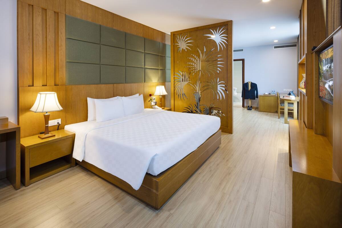 Une chambre d’hôtel avec un grand lit, une tête de lit en bois, deux tables de chevet avec des lampes, un bureau en bois et un panneau décoratif orné de motifs de palmiers.