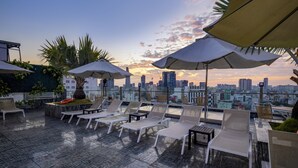 A rooftop terrace with lounge chairs and umbrellas, overlooking a city skyline at sunset.
