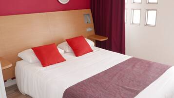 Een hotelkamer met een houten hoofdbord, een bed met witte lakens en rode kussens, een nachtkastje met een lamp en een raam met gordijnen.