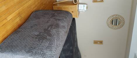Habitación pequeña con cama, una mesita de madera, espejo en la pared y toma de corriente.