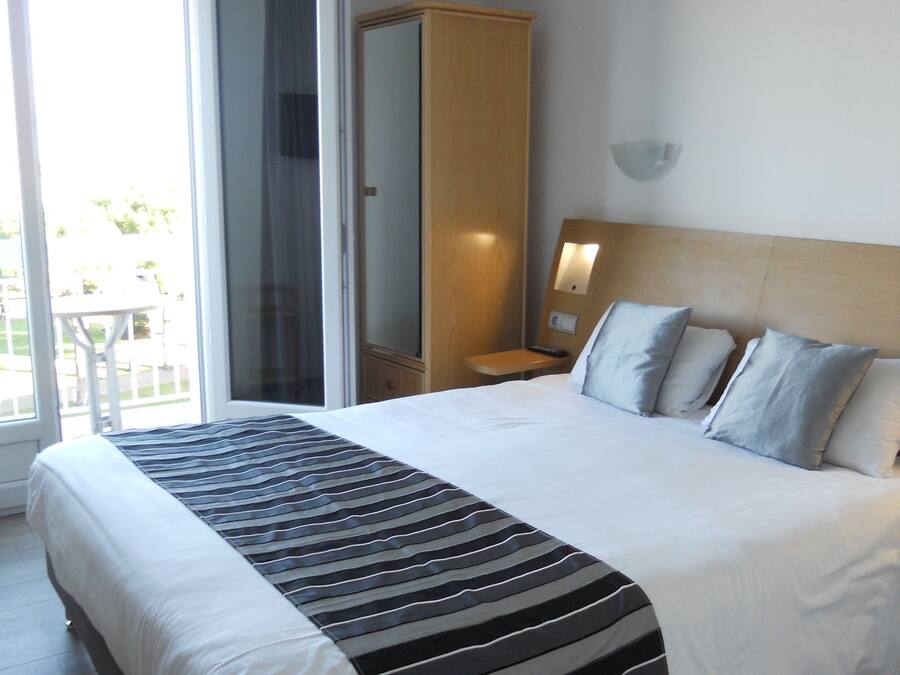 Habitación de hotel con una cama grande, un armario, una mesita de noche con lámpara y una puerta corrediza de vidrio al balcón.