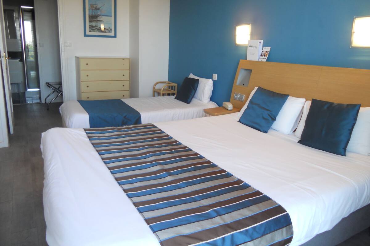 Quarto de hotel com duas camas, uma cabeceira de madeira, uma mesa de cabeceira, uma luminária e um quadro na parede.