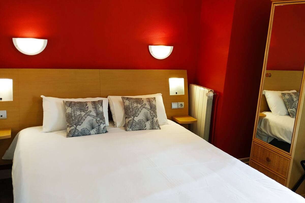 Quarto de hotel com cama grande, duas mesas de cabeceira, um criado-mudo e paredes vermelhas.