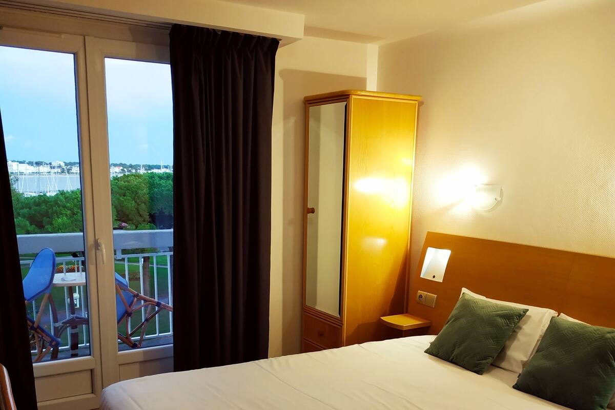 Quarto de hotel com cama, mesa de cabeceira, espelho, janela com cortinas e vista para uma ponte e vegetação lá fora.