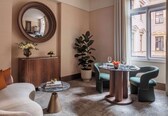 A modern hotel room with a round mirror, a wooden cabinet, a potted plant, two armchairs, a coffee table, and a view of the building outside.