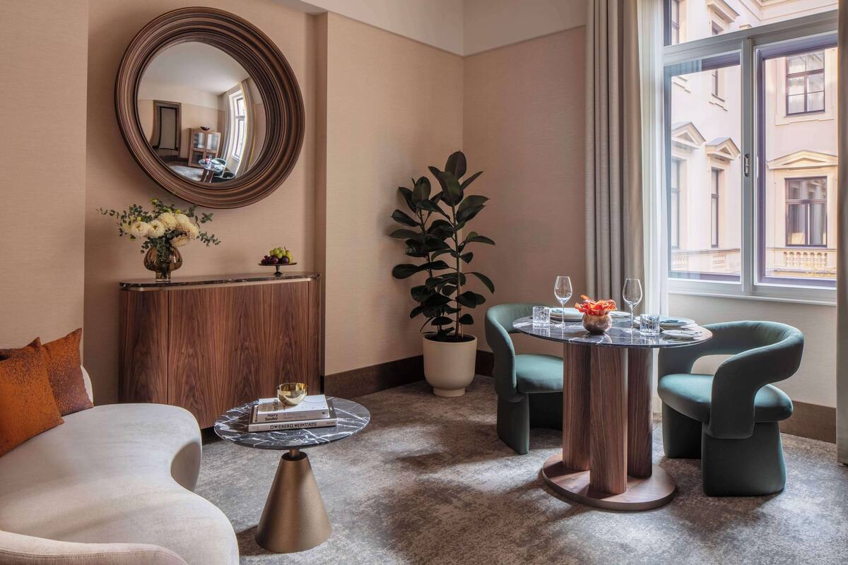Ein modernes Hotelzimmer mit einem runden Spiegel, einem Holzschrank, einer Topfpflanze, zwei Sesseln, einem Couchtisch und Blick auf das Gebäude gegenüber.