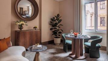 Quarto de hotel moderno com um espelho redondo, um armário de madeira, um vaso de planta, duas poltronas, uma mesa de centro e vista para o edifício lá fora.