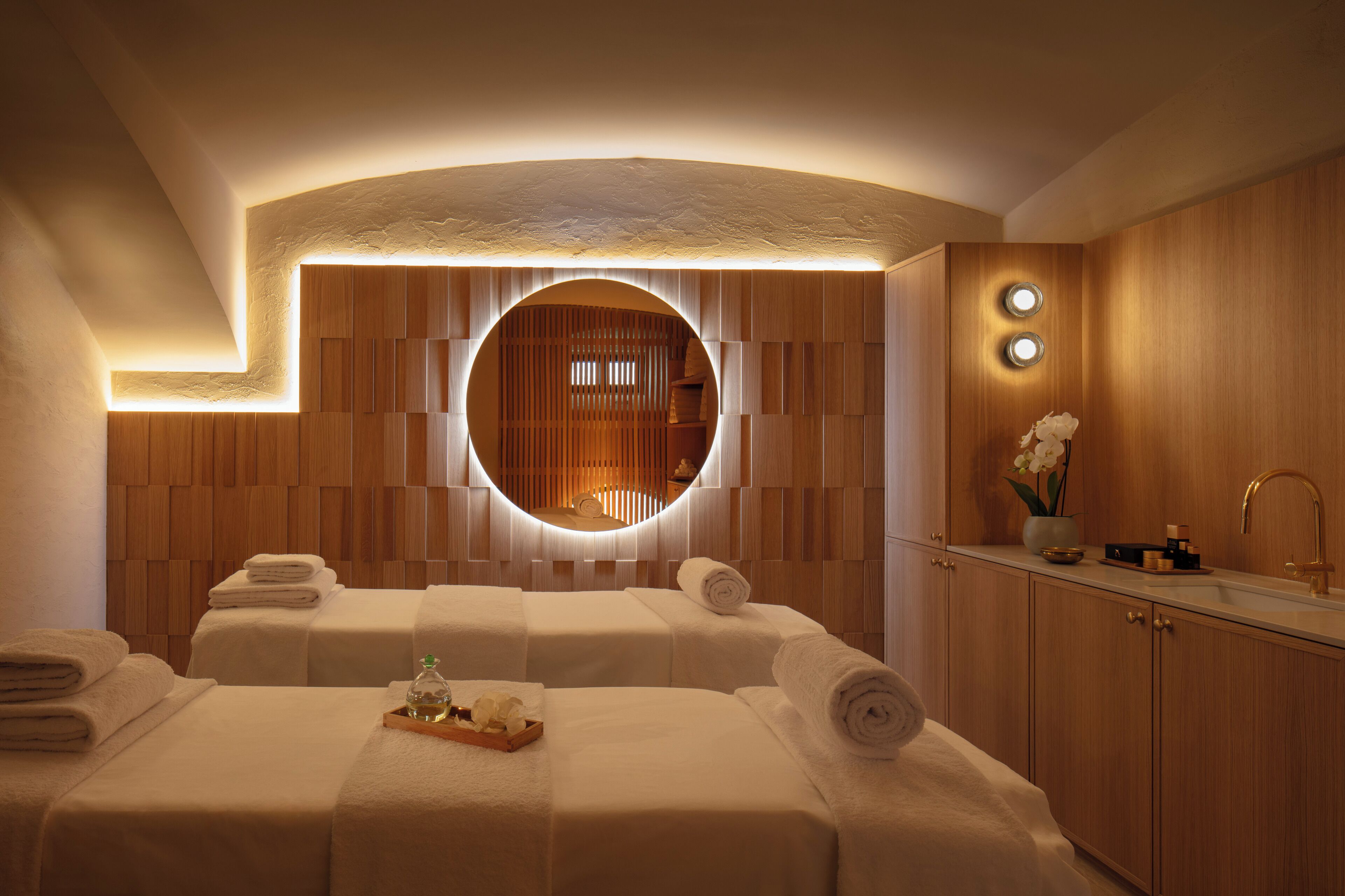 A spa room with two beds, a circular mirror, wooden paneling, and a sink with a vase of flowers.