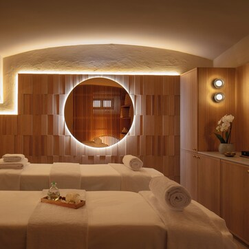 A spa room with two beds, a circular mirror, wooden paneling, and a sink with a vase of flowers.