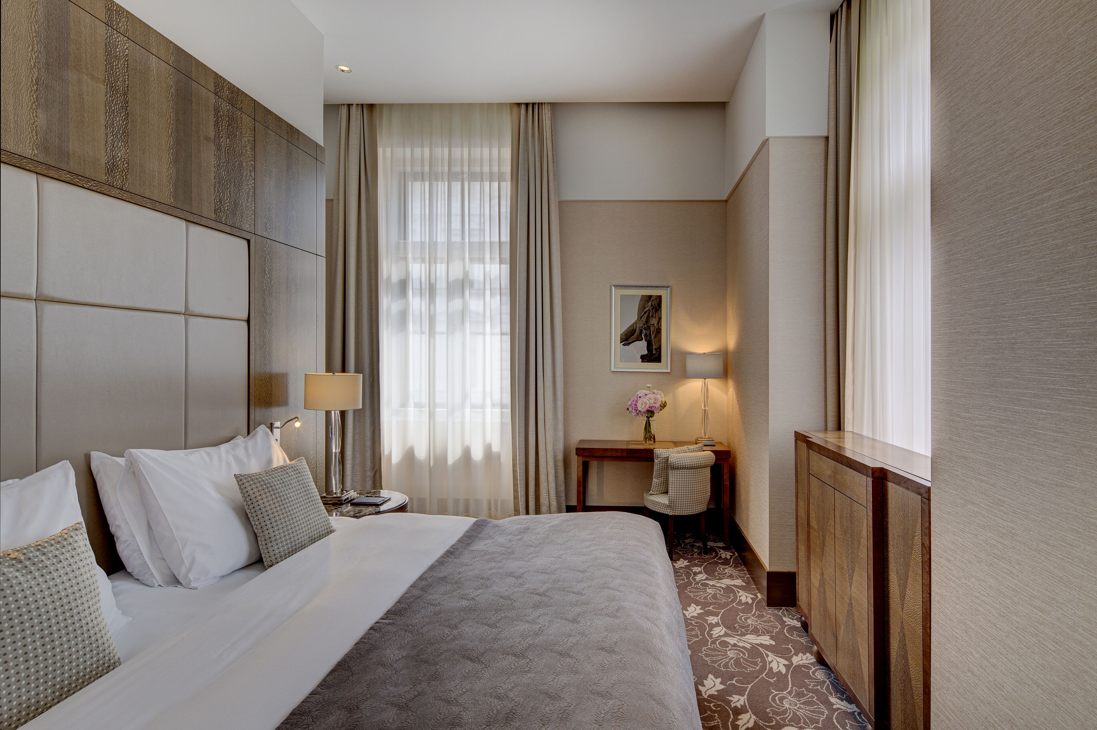 A hotel room with a large bed, a desk with a lamp, a chair, and a small table with a vase.