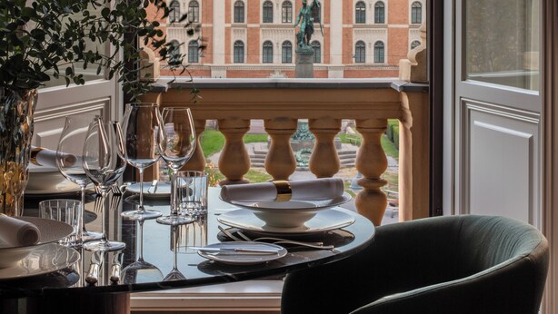 A dining area with a view of a historic building, a balcony with a balustrade, and a table set with wine glasses and plates. - Anantara Palais Hansen Vienna – A Leading Hotel of the World (Vienna)