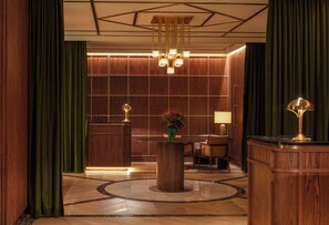 A lobby with a reception desk, a round table with chairs, a vase with flowers, and a lamp.