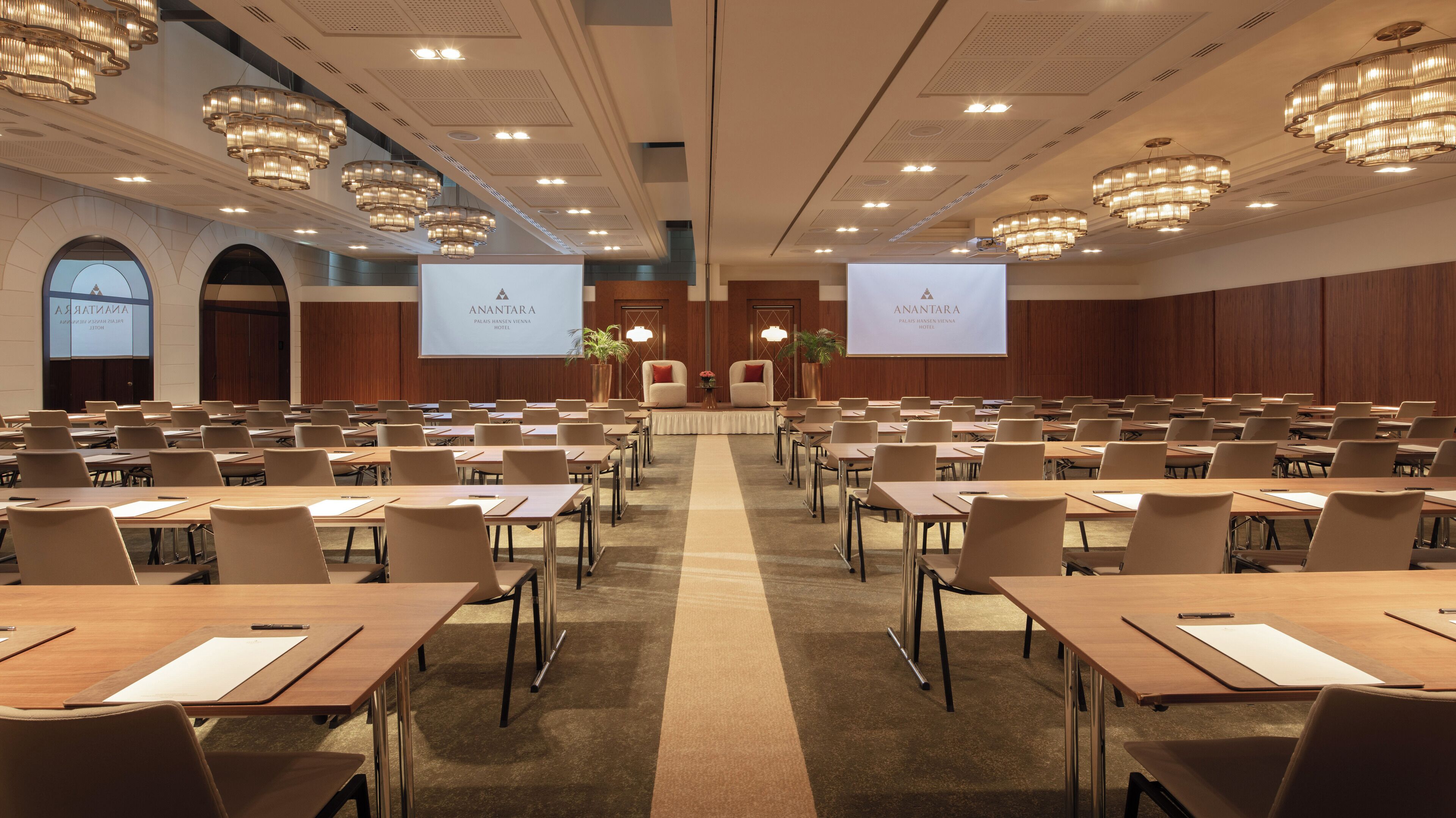 Un salón de conferencias con filas de mesas y sillas, un escenario con un atril y dos pantallas de proyección grandes que muestran el nombre del hotel.