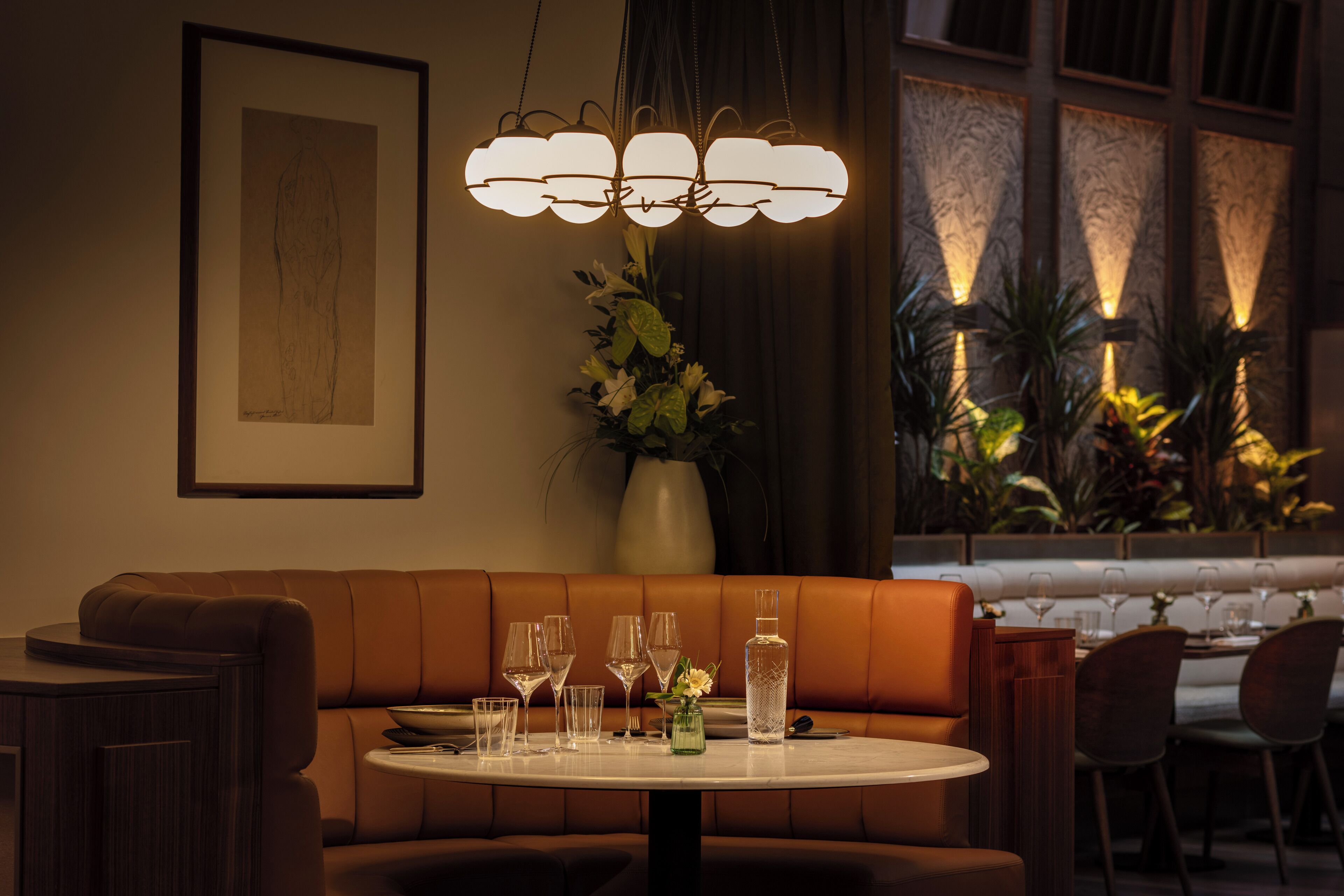 A dining area with a round table, brown leather booth seating, a vase with flowers, and a framed artwork on the wall.