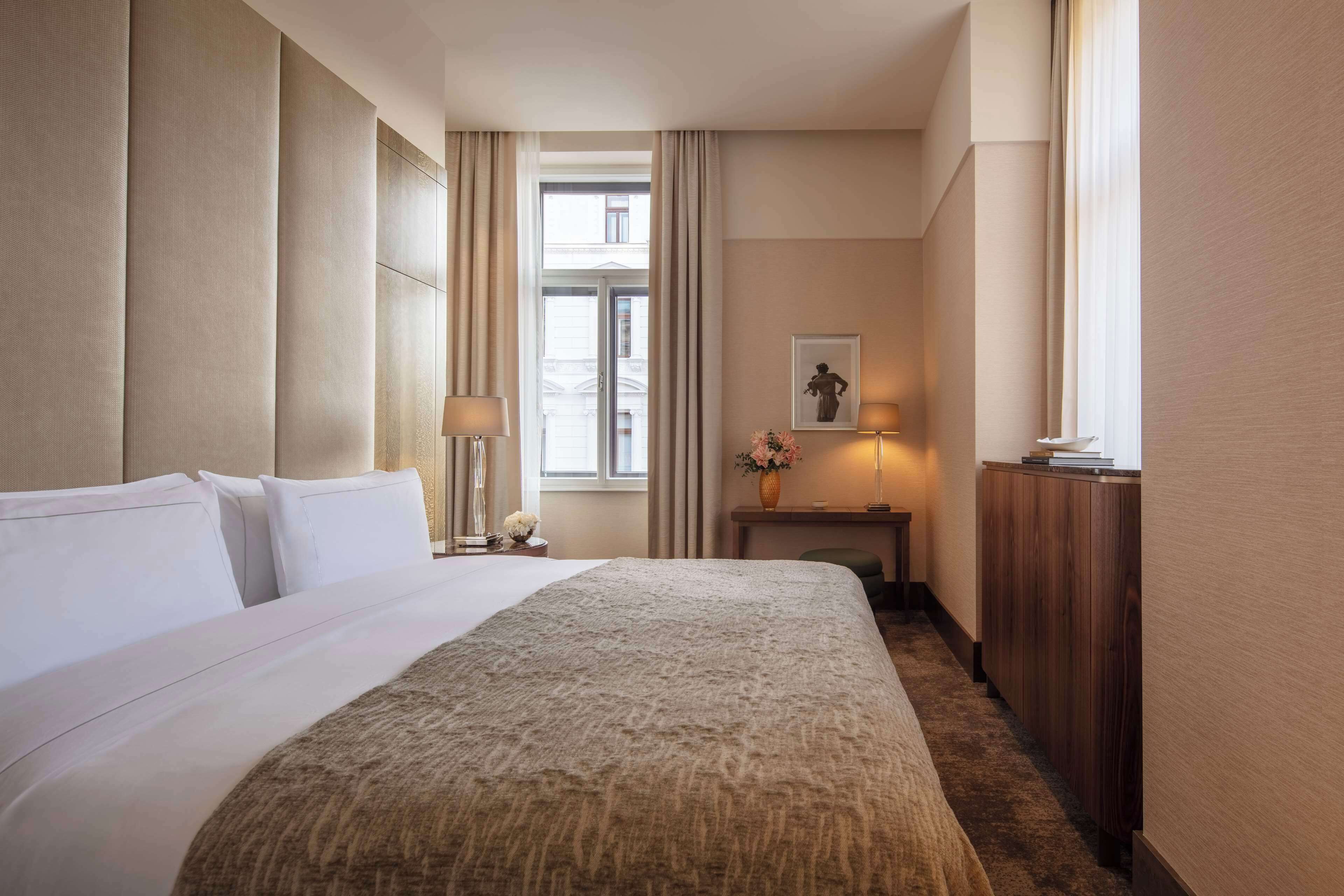 Quarto de hotel com uma cama grande, mesa de cabeceira com abajur, pequena mesa com um vaso e janela com cortinas.