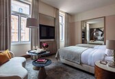 A hotel room with a bed, a sofa, a TV, a small table with a vase and books, and a large mirror.