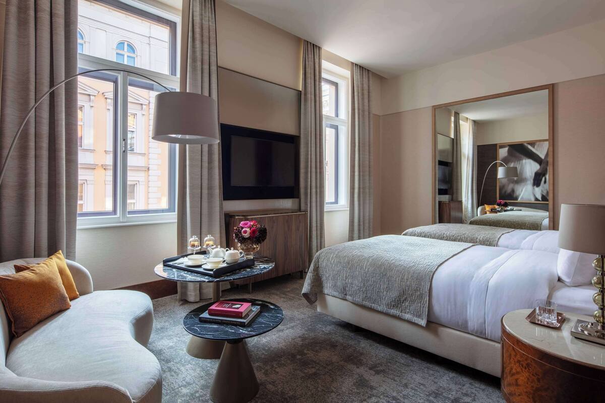 Ein Hotelzimmer mit Bett, Sofa, Fernseher, kleinem Tisch mit Vase und Büchern sowie einem großen Spiegel.