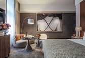 A modern hotel room with a bed, a round table, two armchairs, a coffee table, and a large painting on the wall.