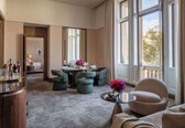 A spacious hotel room with a large dining table set for four, a comfortable seating area with a sofa and armchairs, and a view of a balcony with a railing.