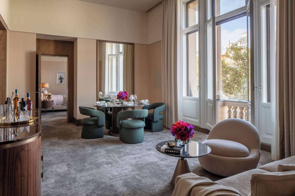 Ein geräumiges Hotelzimmer mit einem großen Esstisch, gedeckt für vier Personen, einer gemütlichen Sitzecke mit Sofa und Sesseln sowie einem Balkon mit Geländer.