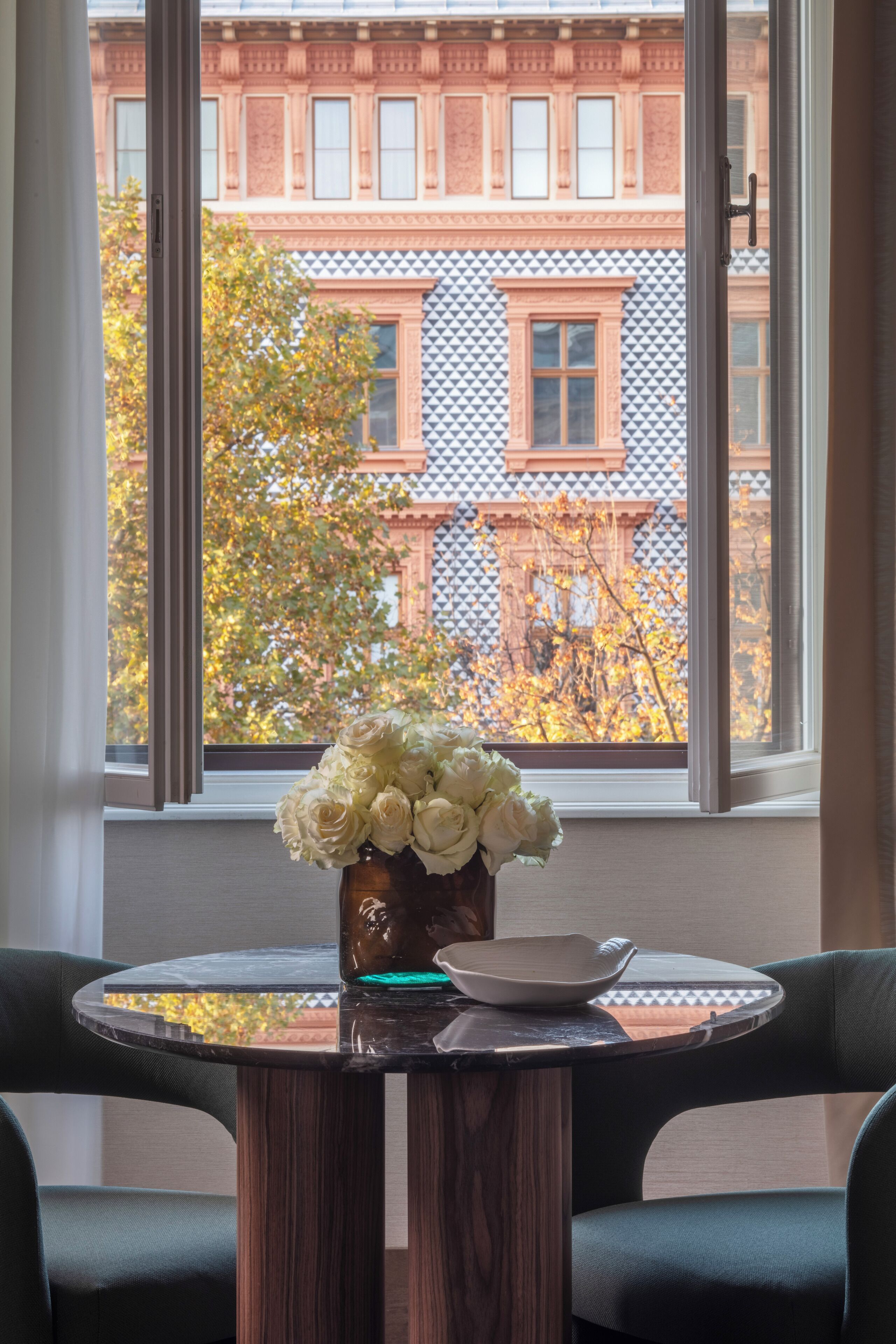 A room with a table, a vase with flowers, and a view of buildings through a window.