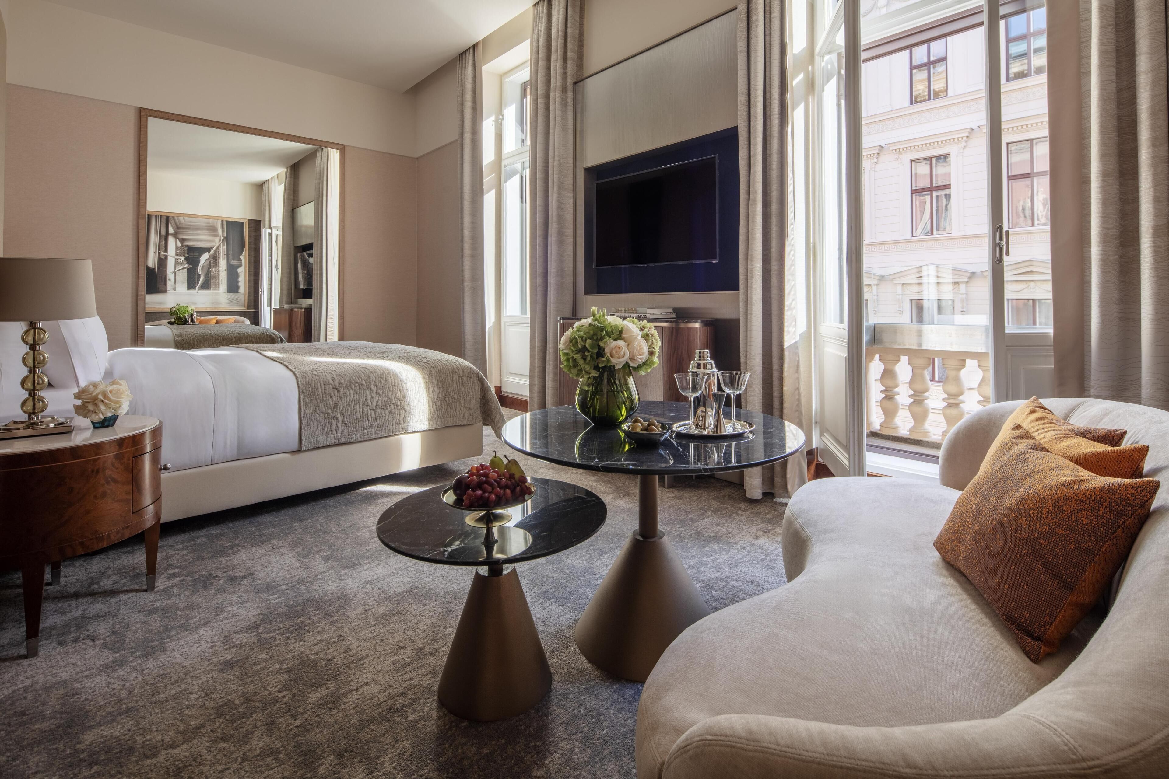 A hotel room with a bed, a sofa, a coffee table, a TV, and a balcony with a view of the city.