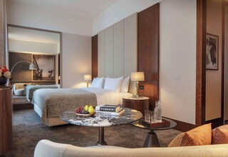 A hotel room with a large bed, two bedside tables with lamps, a round coffee table with fruit and books, and a sofa.