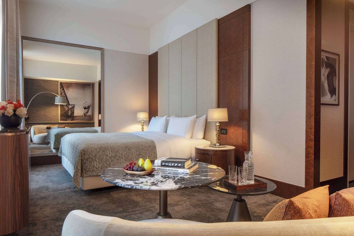Ein Hotelzimmer mit einem großen Bett, zwei Nachttischen mit Lampen, einem runden Couchtisch mit Obst und Büchern sowie einer Couch.