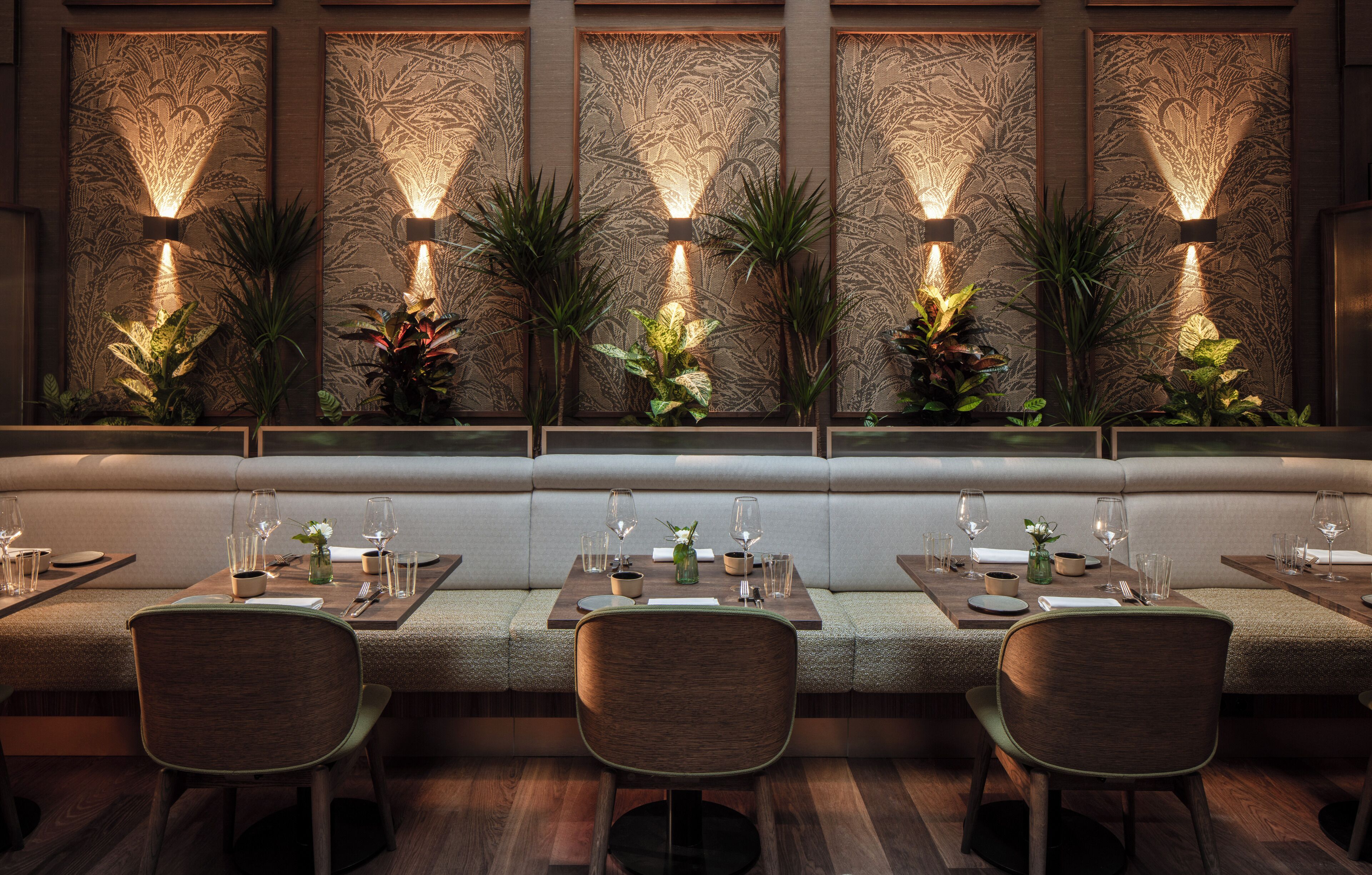 A restaurant with a long bench seating area, tables with chairs, and wall-mounted lighting.