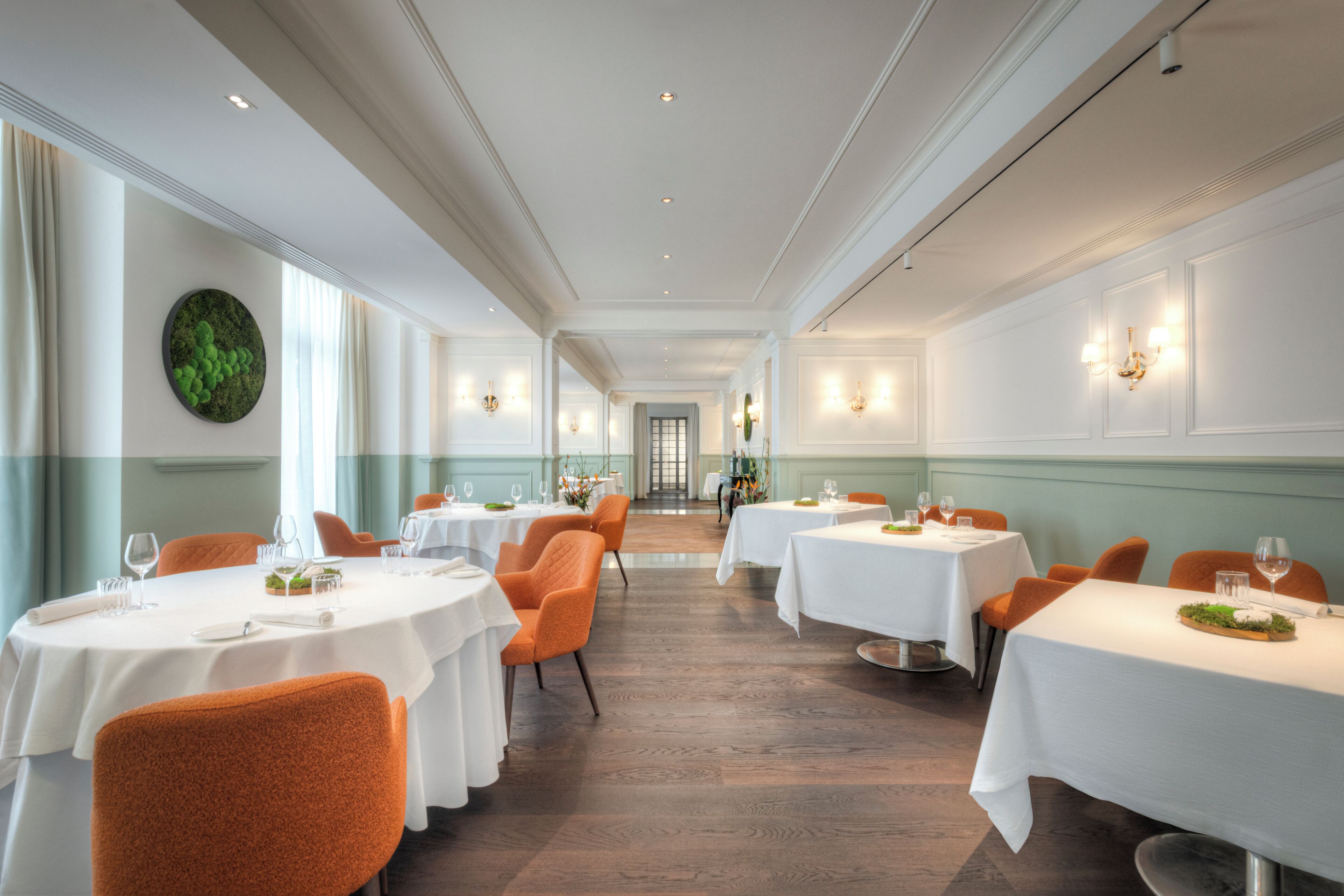 Uma sala de jantar de hotel com mesas redondas, cadeiras laranja e toalhas de mesa brancas.