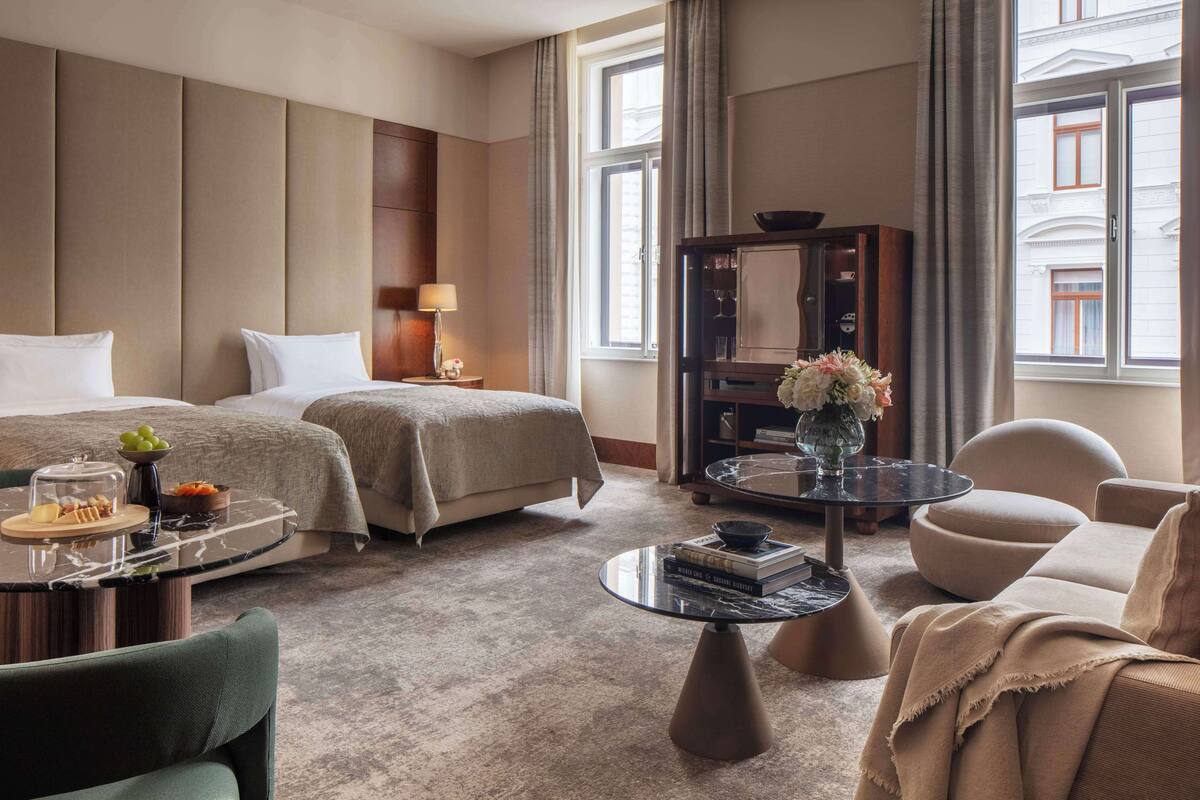 Ein Hotelzimmer mit zwei Betten, einer Sitzecke mit einer Couch und Sesseln, einem kleinen Tisch mit Blumen und Blick auf die Stadt durch große Fenster.