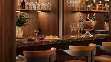 A bar area with a marble countertop, hanging wine glasses, and bottles of champagne. There are chairs and a plant in the background.