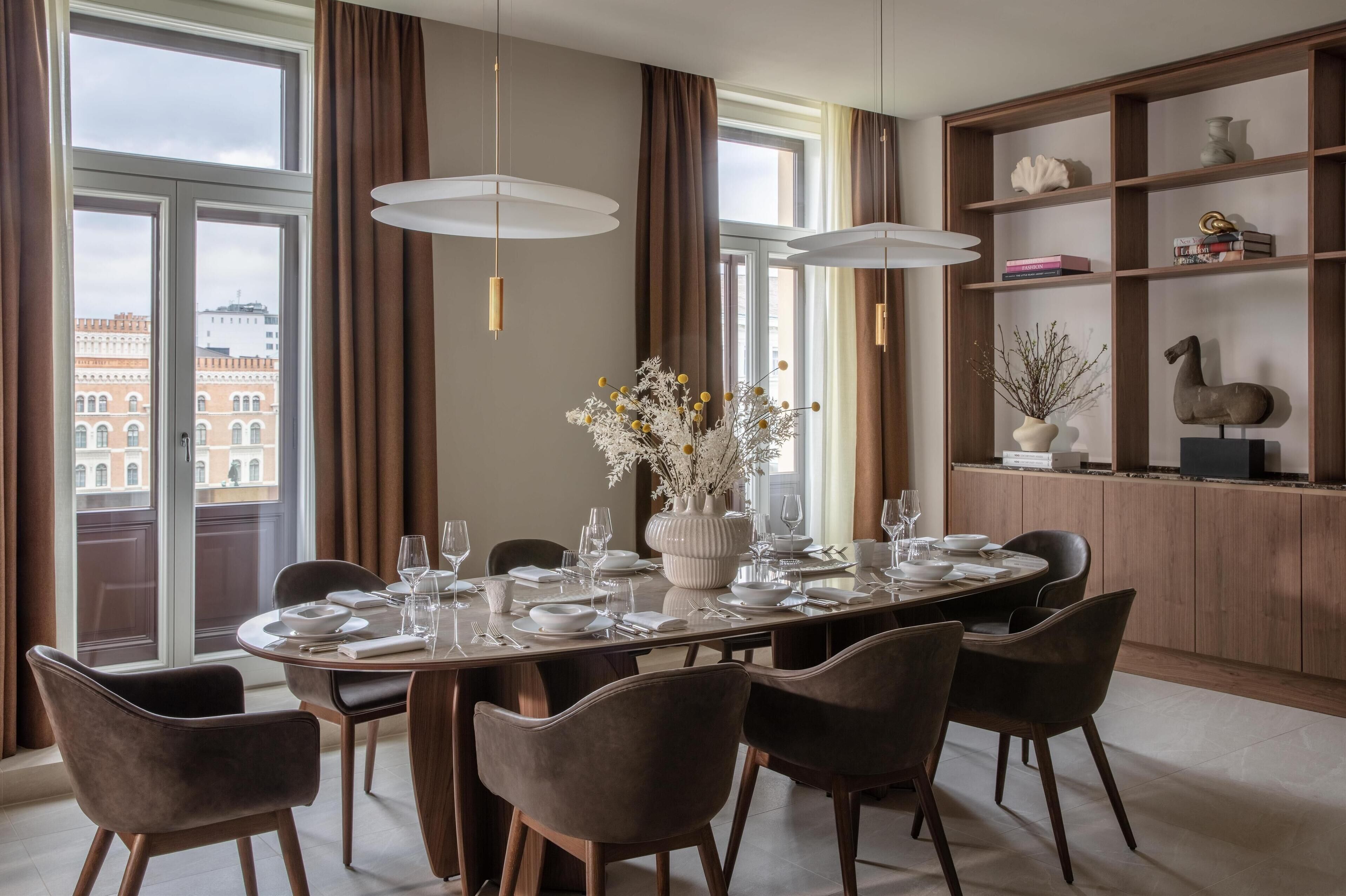 Un comedor con una mesa redonda dispuesta para seis personas, rodeada de seis sillas. Hay un ventanal con cortinas, una estantería con objetos decorativos y un jarrón con flores sobre la mesa.