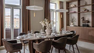 Un comedor con una mesa redonda dispuesta para seis personas, rodeada de seis sillas. Hay un ventanal con cortinas, una estantería con objetos decorativos y un jarrón con flores sobre la mesa.