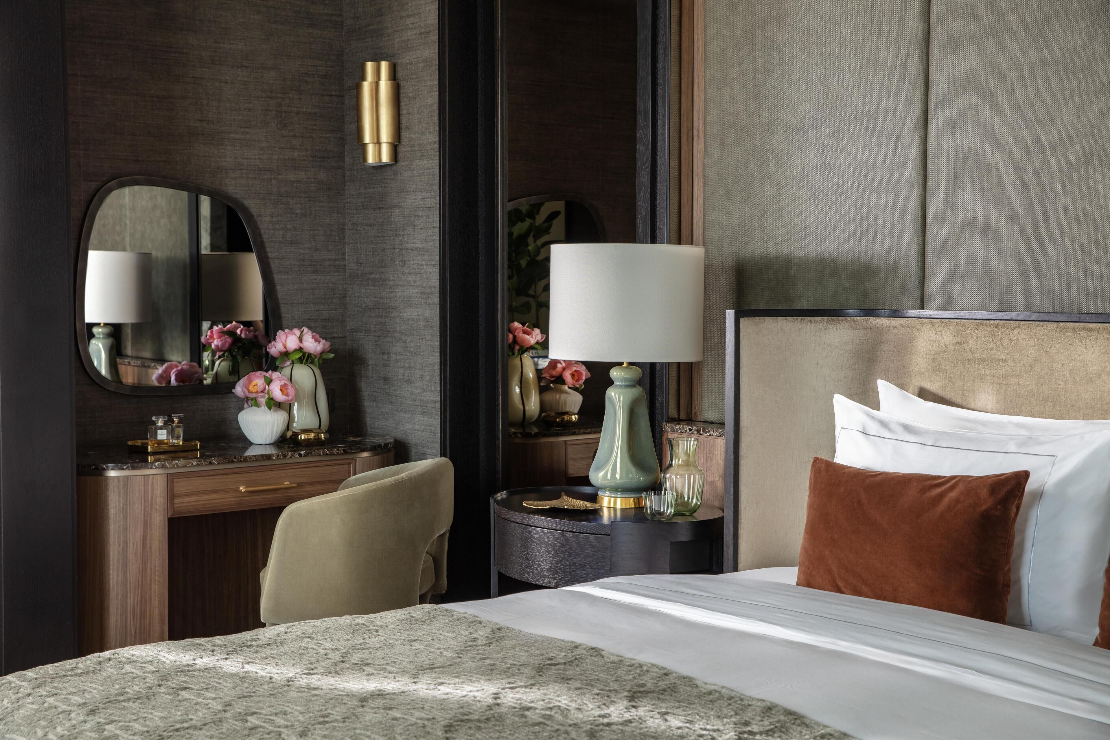 Quarto de hotel com uma cama, uma mesa com cadeira, uma cômoda com espelho e uma mesa de cabeceira com abajur.