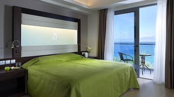 A hotel room with a large bed, a nightstand, a lamp, and a view of the sea through a balcony door.