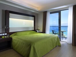 Habitación de hotel con una cama grande, una mesita de noche, una lámpara y vista al mar a través de una puerta de balcón.