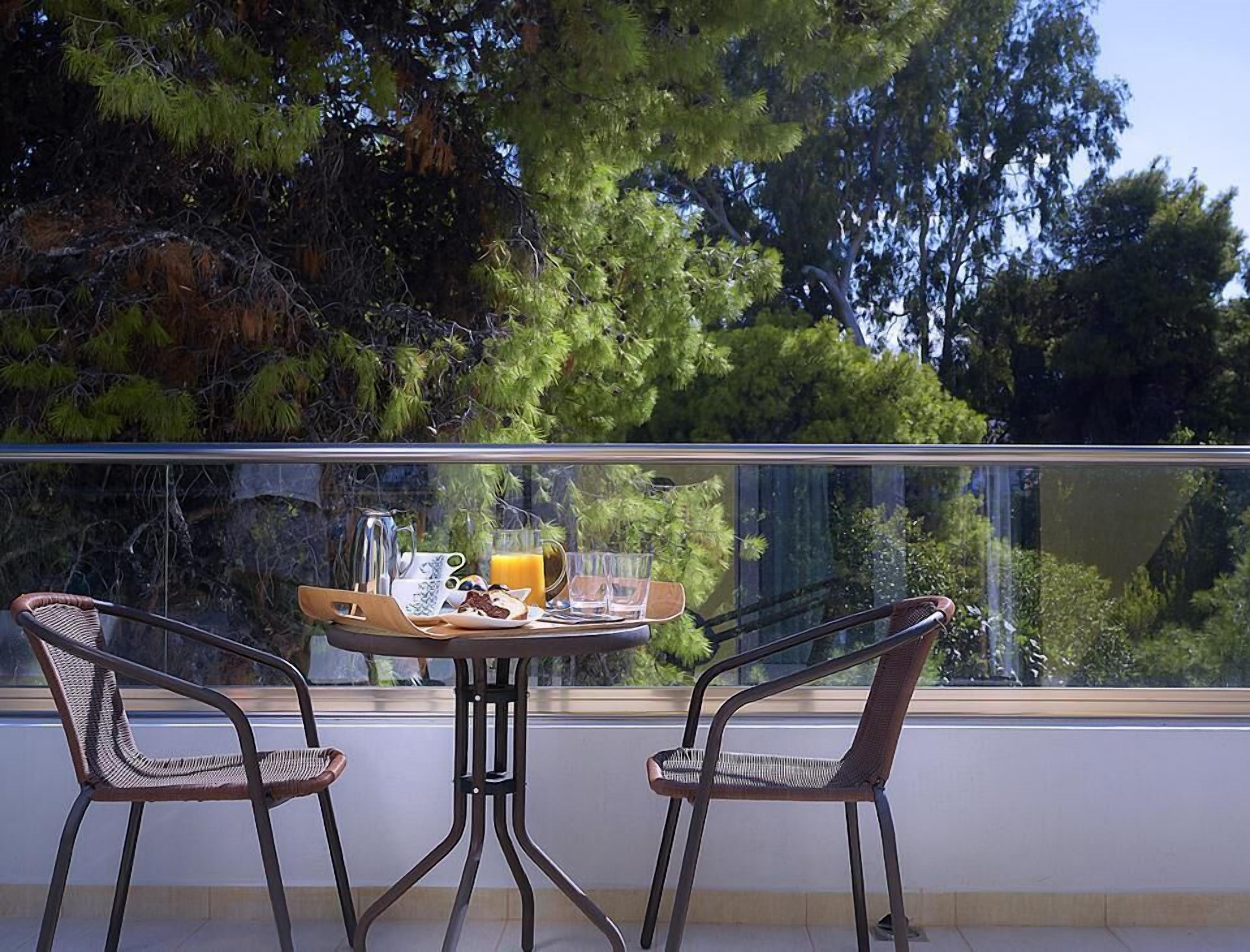 A balcony with two wicker chairs, a small round table with breakfast items, and a view of trees and sky.