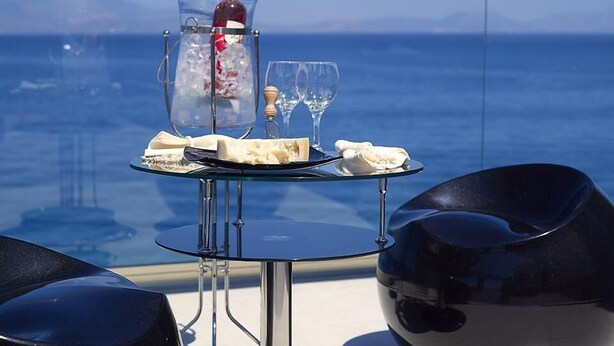 Una mesa con una botella de vino, dos copas y un plato de queso y galletas, frente a un mar azul y montañas a lo lejos.